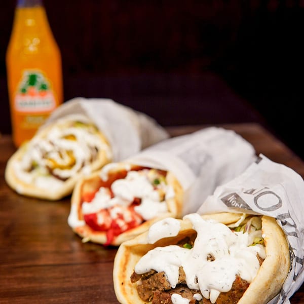 Three Haazs halal gyros with white sauce and a bottle of orange soda on a wooden table.