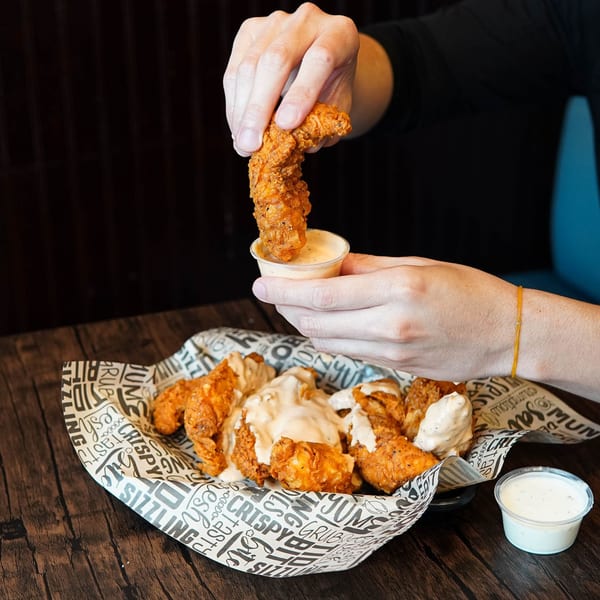 A hand dips a crispy Haazs chicken tender into a small cup of sauce, with more tenders in a basket.