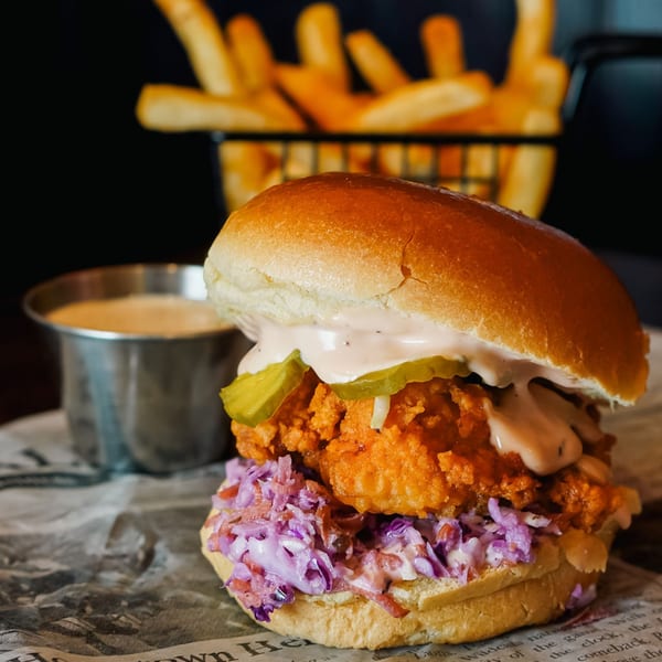 A Haazs fried chicken sandwich with coleslaw, pickles, and sauce, with fries and dip in the background.
