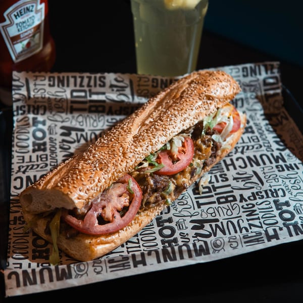 A Haazs halal cheesesteak sandwich with tomatoes and lettuce on branded paper, with ketchup and a drink.